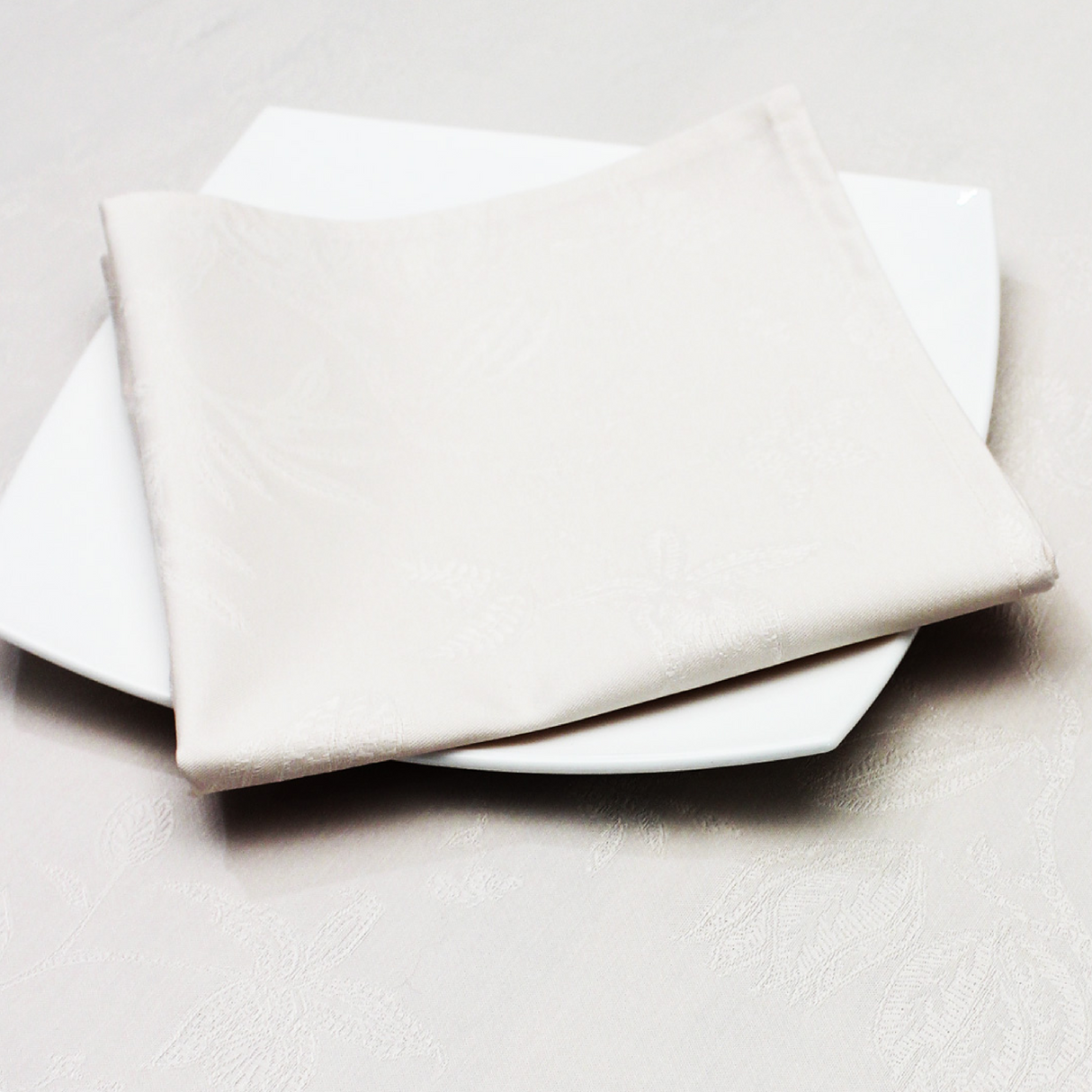 A folded beige Christmas napkin with a damask pattern, placed on a white plate with a similar patterned tablecloth in the background.