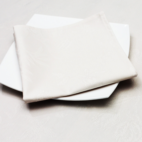A folded beige Christmas napkin with a damask pattern, placed on a white plate with a similar patterned tablecloth in the background.