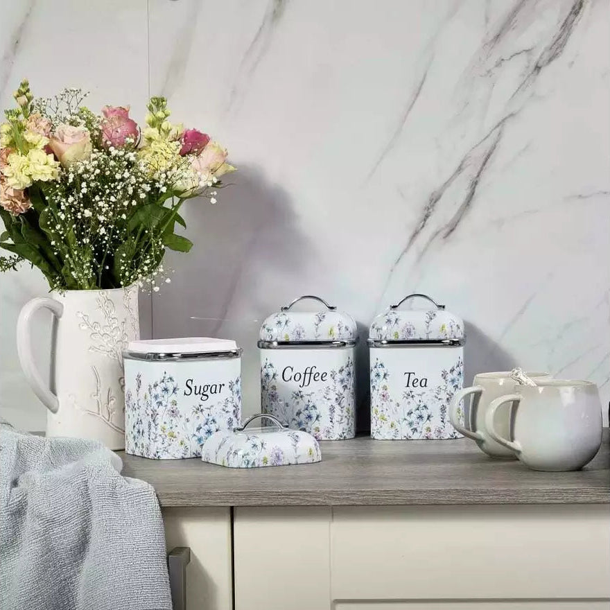 A set of three floral-patterned metal canisters labeled Sugar, Coffee, and Tea, styled on a kitchen counter next to a mug, a vase of flowers, and a towel against a marble wall.