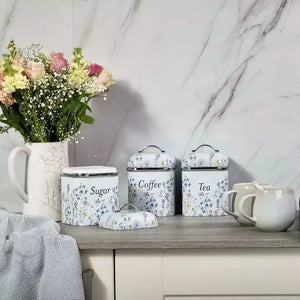 A set of three floral-patterned metal canisters labeled Sugar, Coffee, and Tea, styled on a kitchen counter next to a mug, a vase of flowers, and a towel against a marble wall.